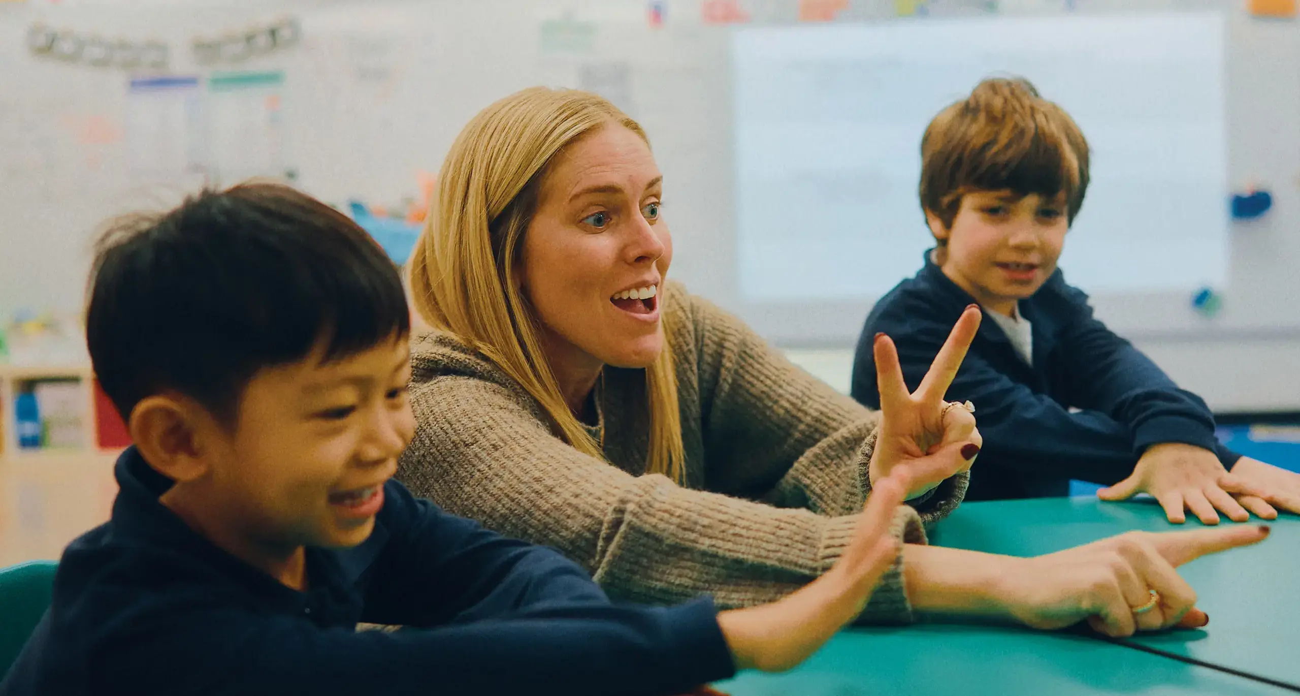 A teacher and two young students are sitting at a table in a classroom. The teacher is speaking, holding two fingers up, while the students smile.