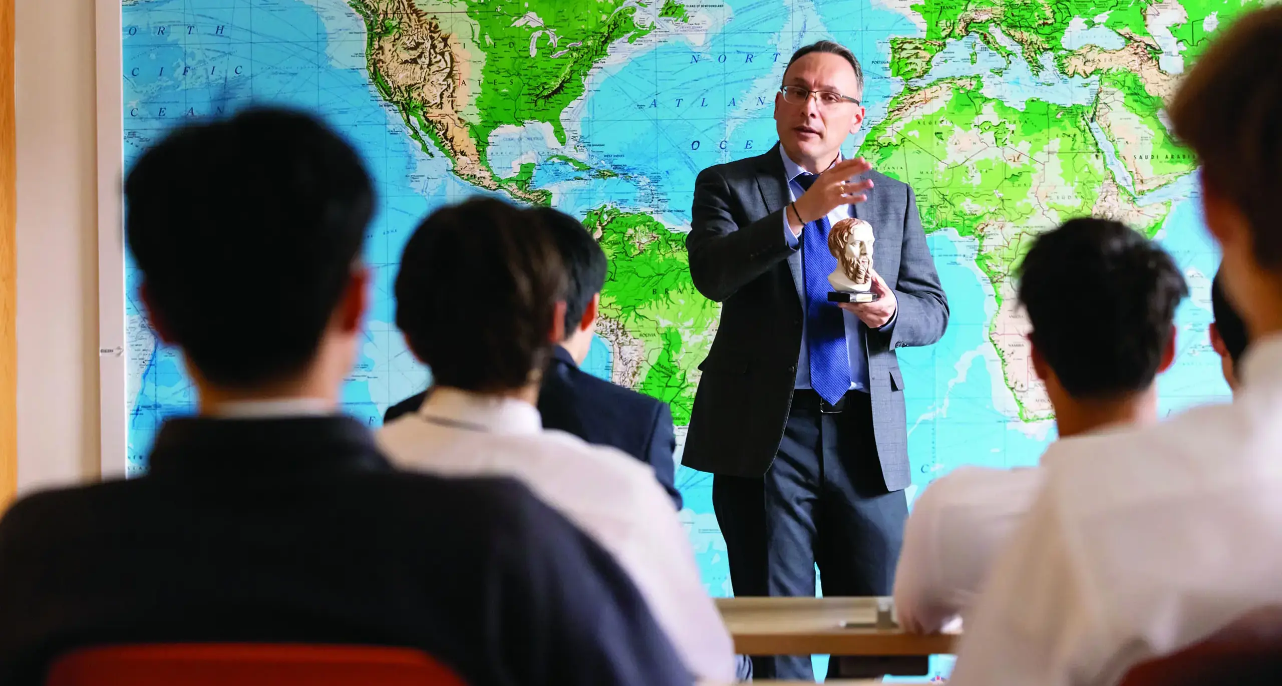 A teacher lectures in front of a world map, holding a small bust, with students seated in front of him.