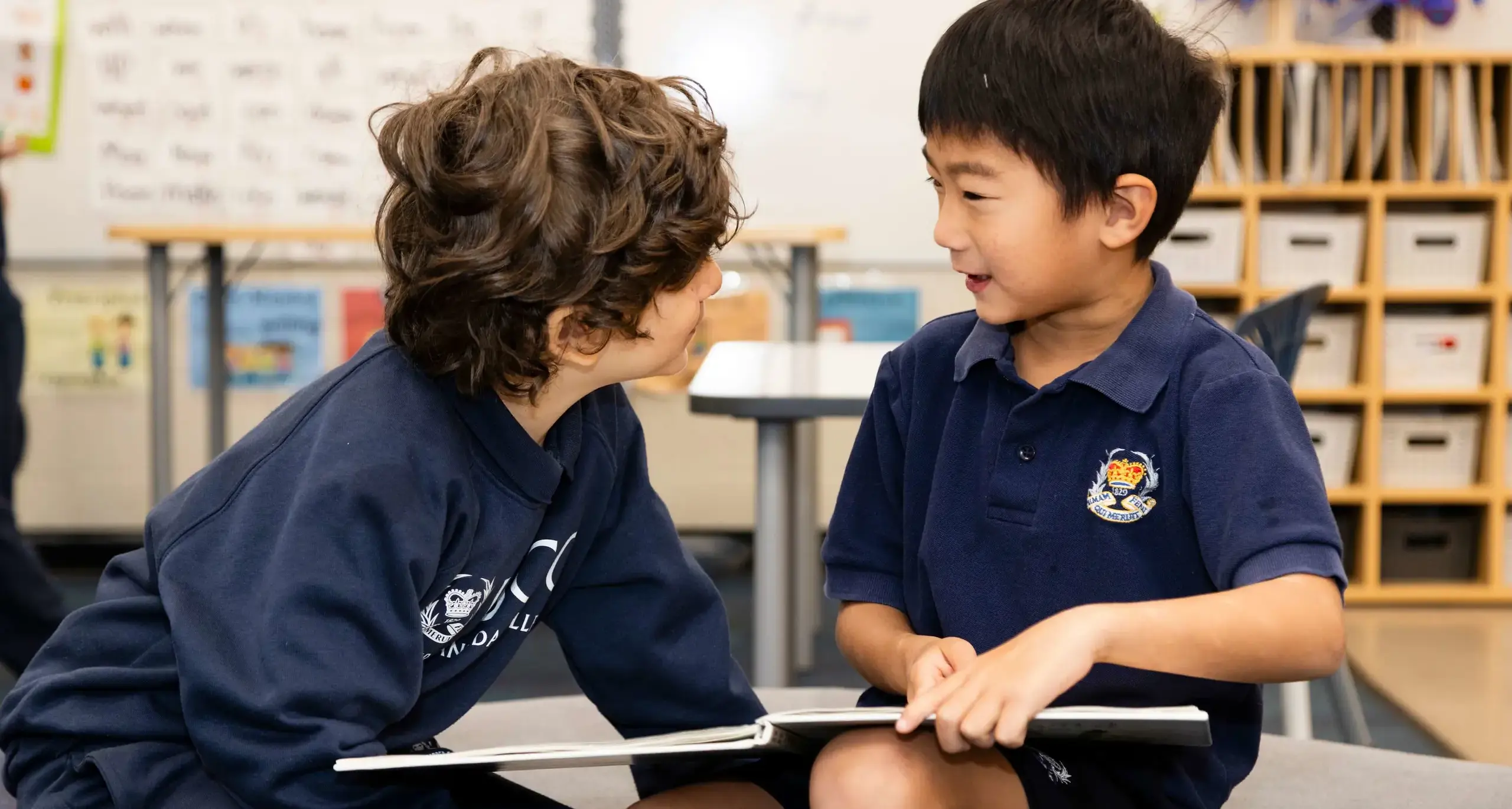 Two young students in navy school uniforms sit together, sharing a book and engaging in a friendly conversation.